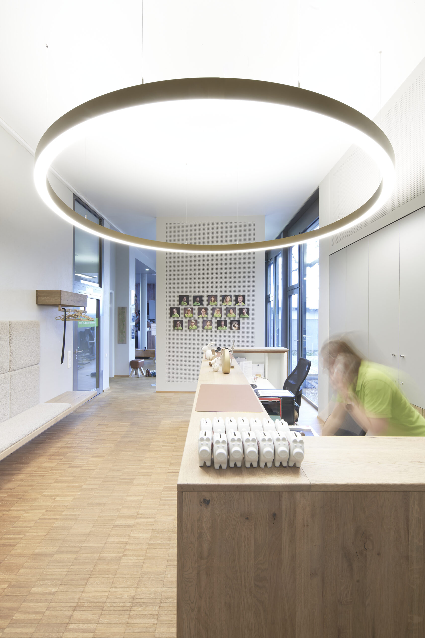 Innenansicht Praxis für Physiotherapie, Eingangsbereich mit Theke und riesiger goldenen Ringleuchte. Parkettboden und passende Oberflächen in dezentem grau und weiß geölter Eiche