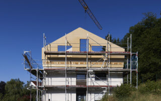 Aufstockung eines bestehenden Wohnhauses mit einer kompletten Wohnung. Aufrichtung der vorgefertigten Holzkonstruktion, Brettstapeldecke, Holz-Ständerwände, noch alles im rohen Zustand. Kran und blauer Himmel bei Sonnenschein