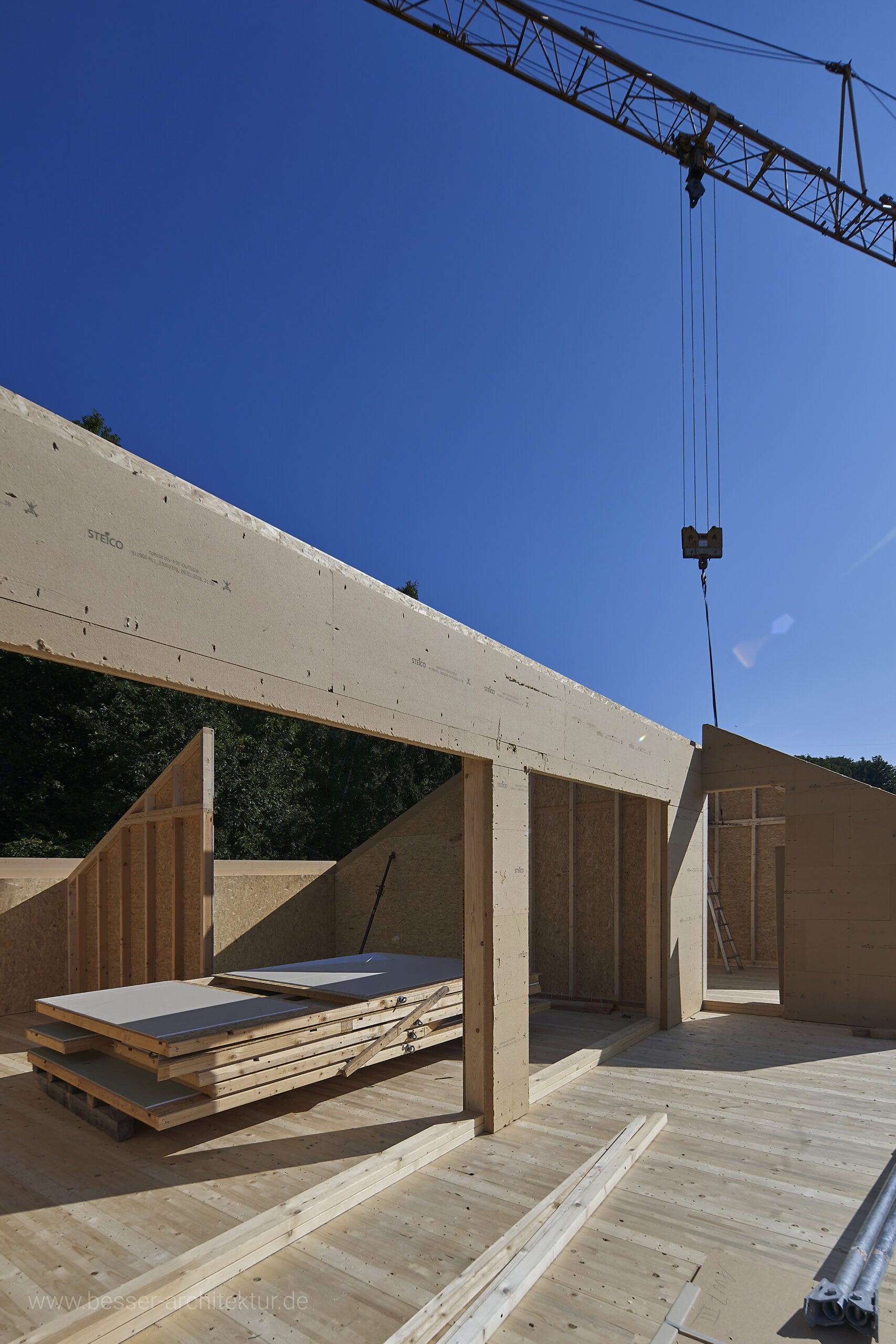 Aufstockung eines bestehenden Wohnhauses mit einer kompletten Wohnung. Aufrichtung der vorgefertigten Holzkonstruktion, Brettstapeldecke, Holz-Ständerwände, noch alles im rohen Zustand. Kran und blauer Himmel bei Sonnenschein 