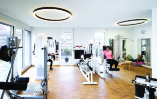 Innenansicht Praxis für Physiotherapie, Trainingsbereich mit Geräten, Ringleuchten an der Decke, Parkettboden und passende Oberflächen in dezentem grau und weiß geölter Eiche, leicht undeutlich 2 Patienten beim Training